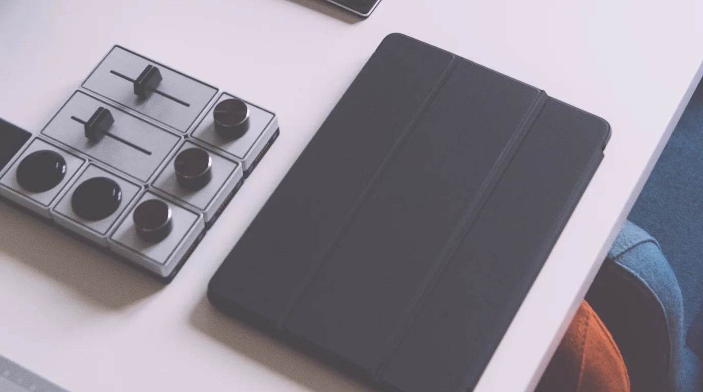 A white desk from above with a tablet in a folding cover and a metal deck of sliders and dials beside it.